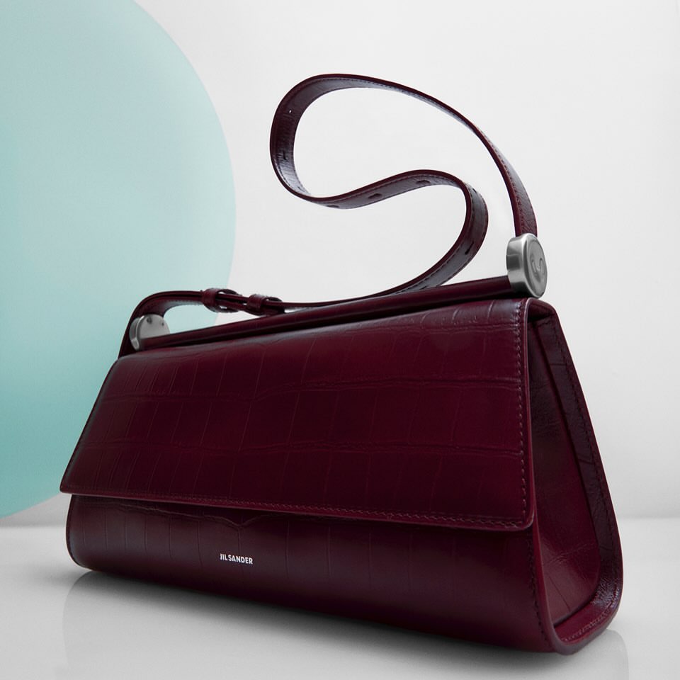 Studio product shot of a burgundy Jil Sander handbag with silver hardware and handle on a white surface against a light blue background.