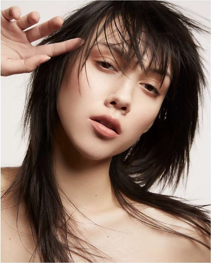 Close-up studio portrait of an Asian woman with dark hair, soft makeup, and hand near her face, set against a bright white background.