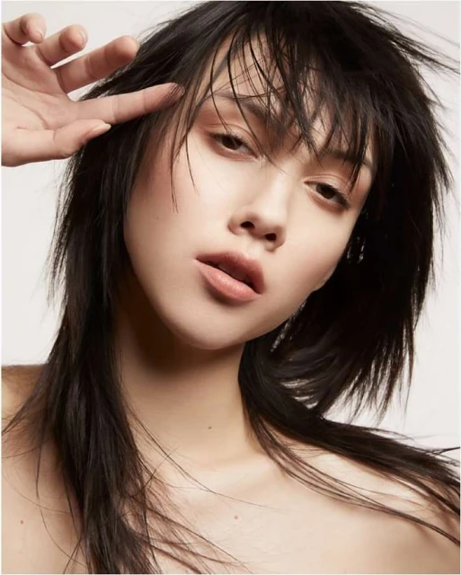 Close-up studio portrait of an Asian woman with dark hair, soft makeup, and hand near her face, set against a bright white background.