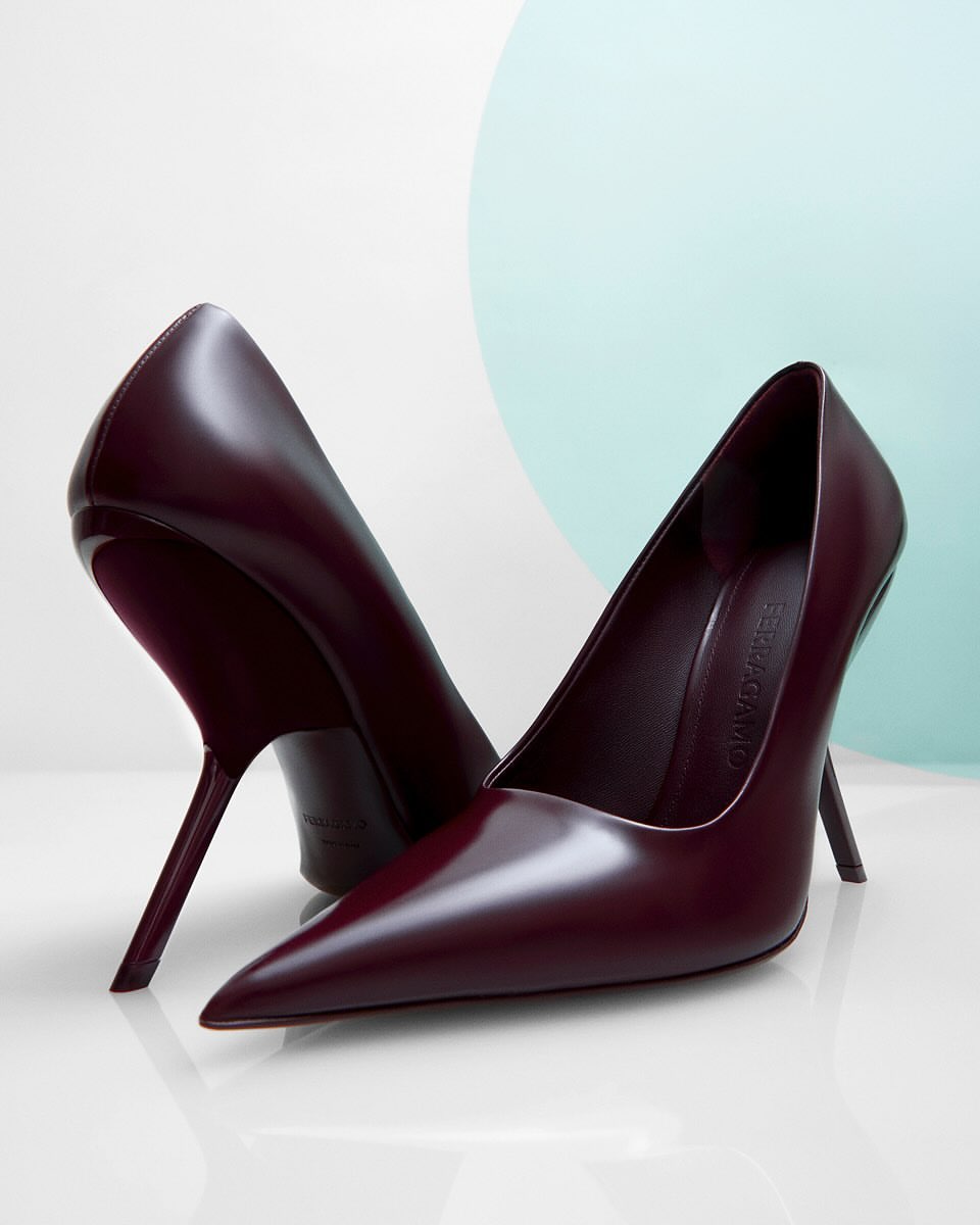 Elegant pointed-toe stiletto heels in deep burgundy on a glossy white surface, with a soft teal and white backdrop, fashion-forward footwear.
