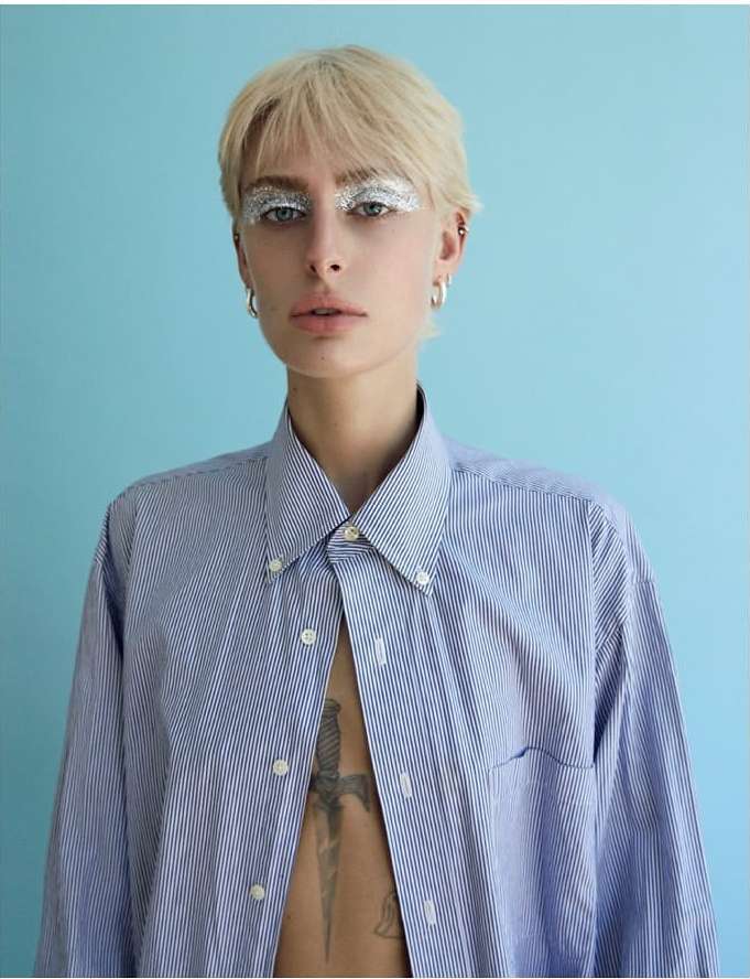 Portrait of a young adult woman with short blond hair, silver eye makeup, and a blue striped shirt against a light blue backdrop.