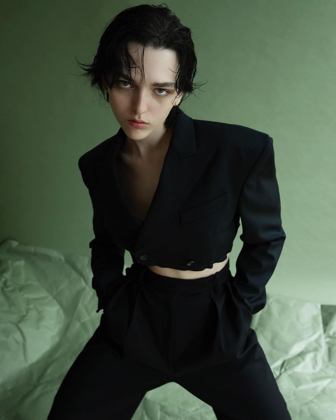 Woman in dark cropped blazer and pants against light green backdrop with crumpled paper. Studio portrait showcasing fashion and beauty.