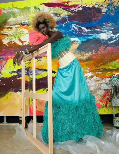 Fashion shot of a black woman with blond hair in a teal feather-trimmed dress, leaning on a wood frame with a painted backdrop.