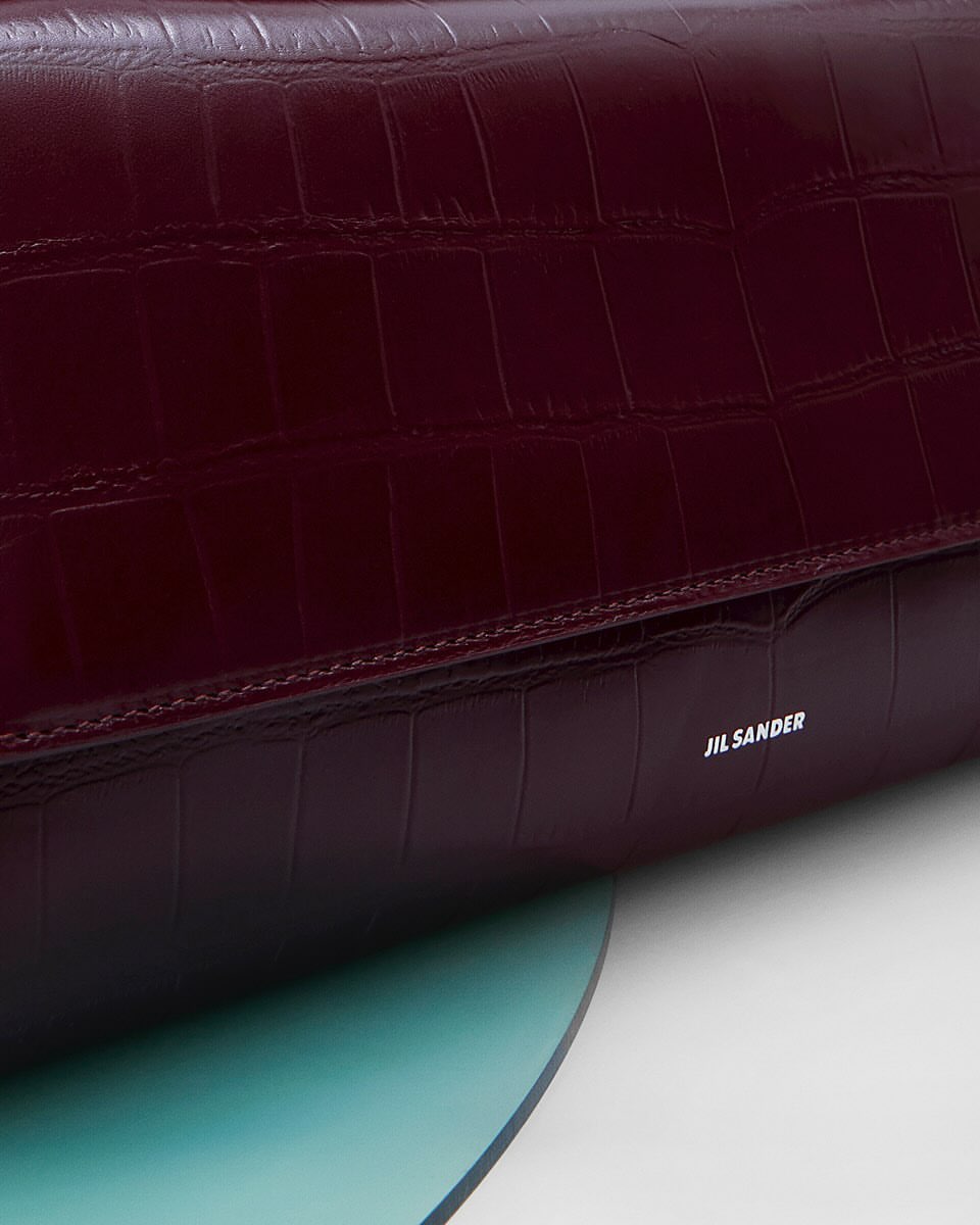 Burgundy crocodile-embossed leather handbag with flap closure, JIL SANDER logo, set against a teal and white background.