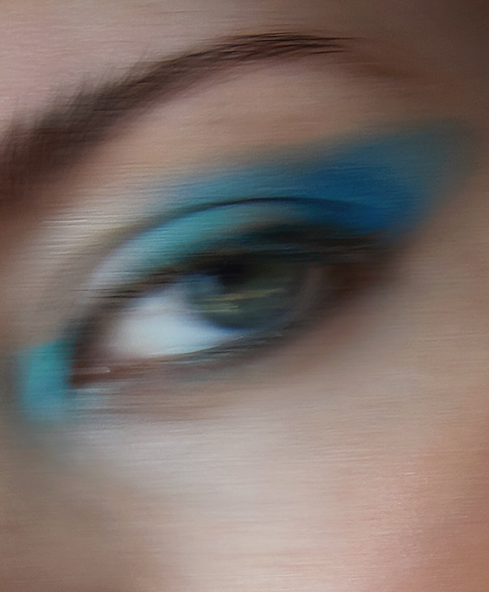 Close-up of a woman's eye with vivid teal eyeshadow extending to the eyebrow and dark brown eyebrow, captured in diffused lighting.