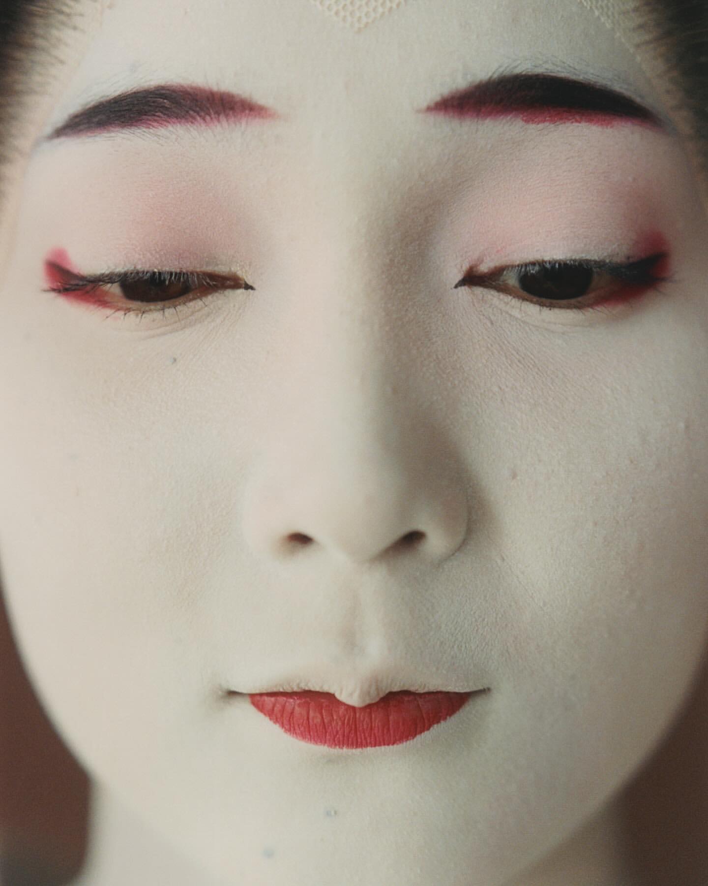 Close-up of a woman with traditional Japanese makeup: white face, red lips, dark eyebrows, and red eyeliner, eyes looking down.