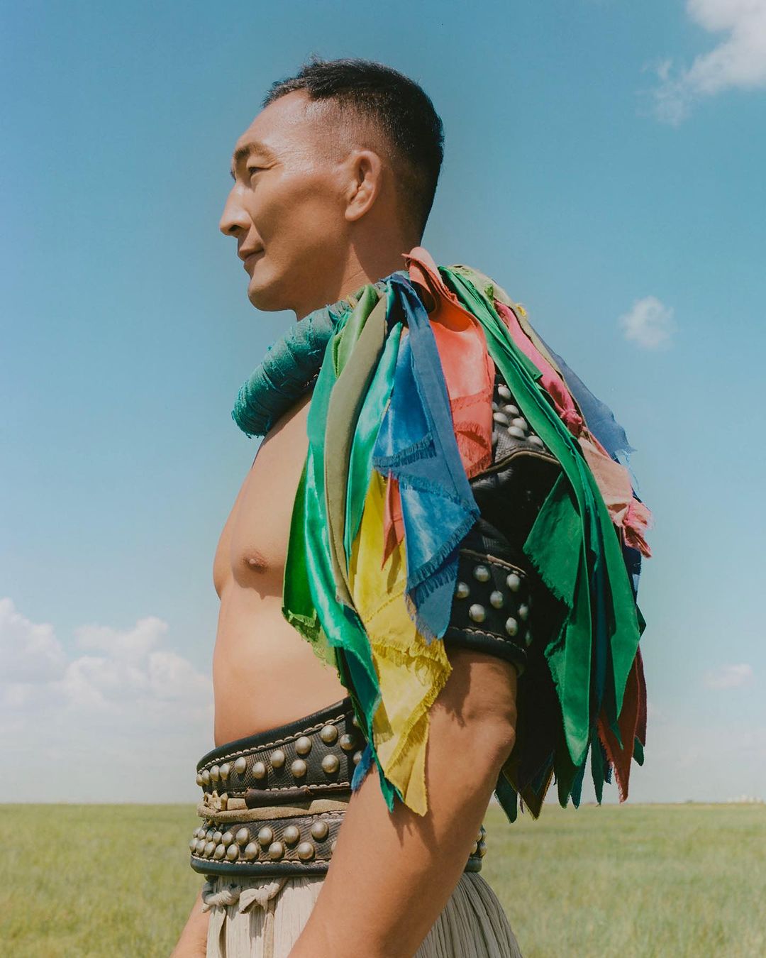 Man in traditional Mongolian clothing with a colorful shoulder piece stands in a grassy field under a blue sky on a sunny day.