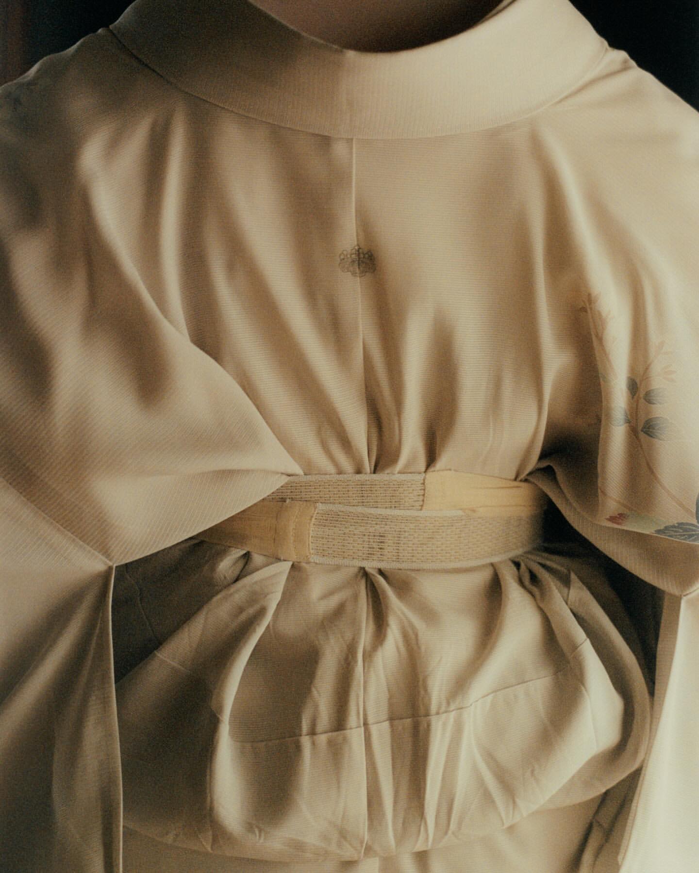 Close-up of a person wearing a tan kimono with a visible obi. The kimono has subtle floral patterns, and the shot is softly lit.