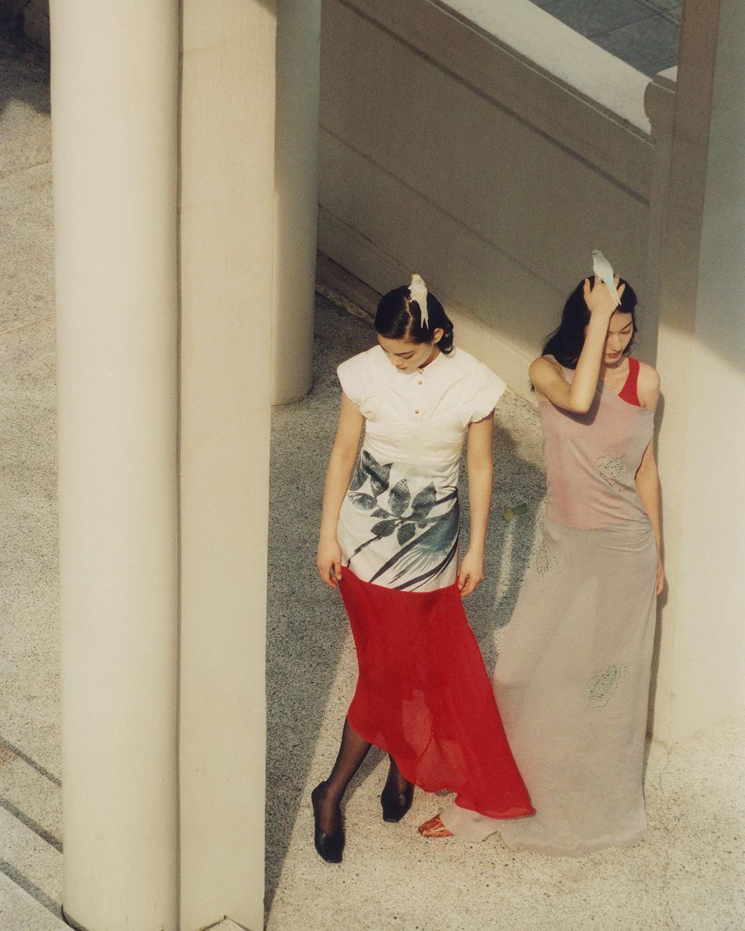 Two women in dresses posing near white pillars. One wears red skirt, the other wears pink. Outdoor fashion photography.
