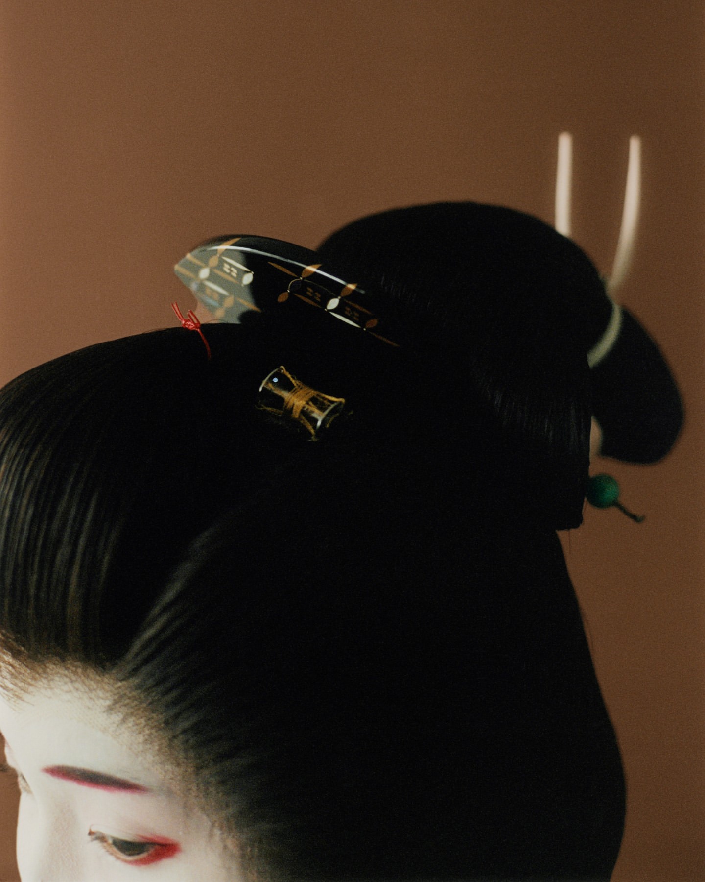 Close-up of a Japanese woman with pale face makeup, wearing black hair styled with ornamental hairpins, against a brown background.