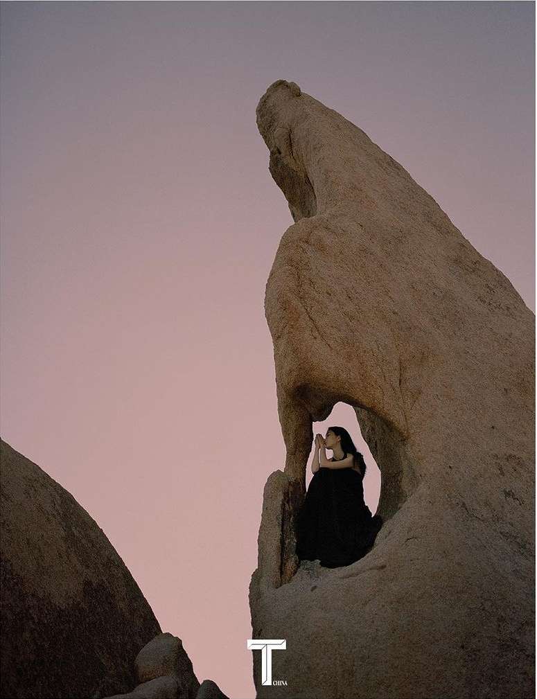 Woman in flowing black dress poses inside a rock formation at dusk with a light purple sky in a landscape scene with warm tones.