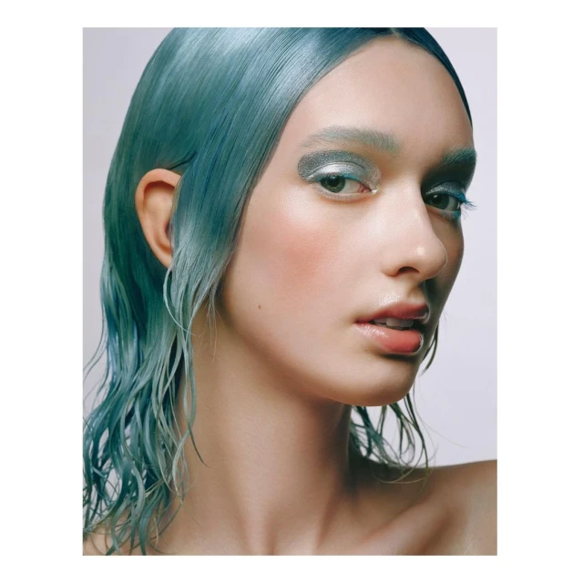 Close-up shot of a female model with blue wet-look hair and silvery eyeshadow posing against a plain backdrop in a studio setting.