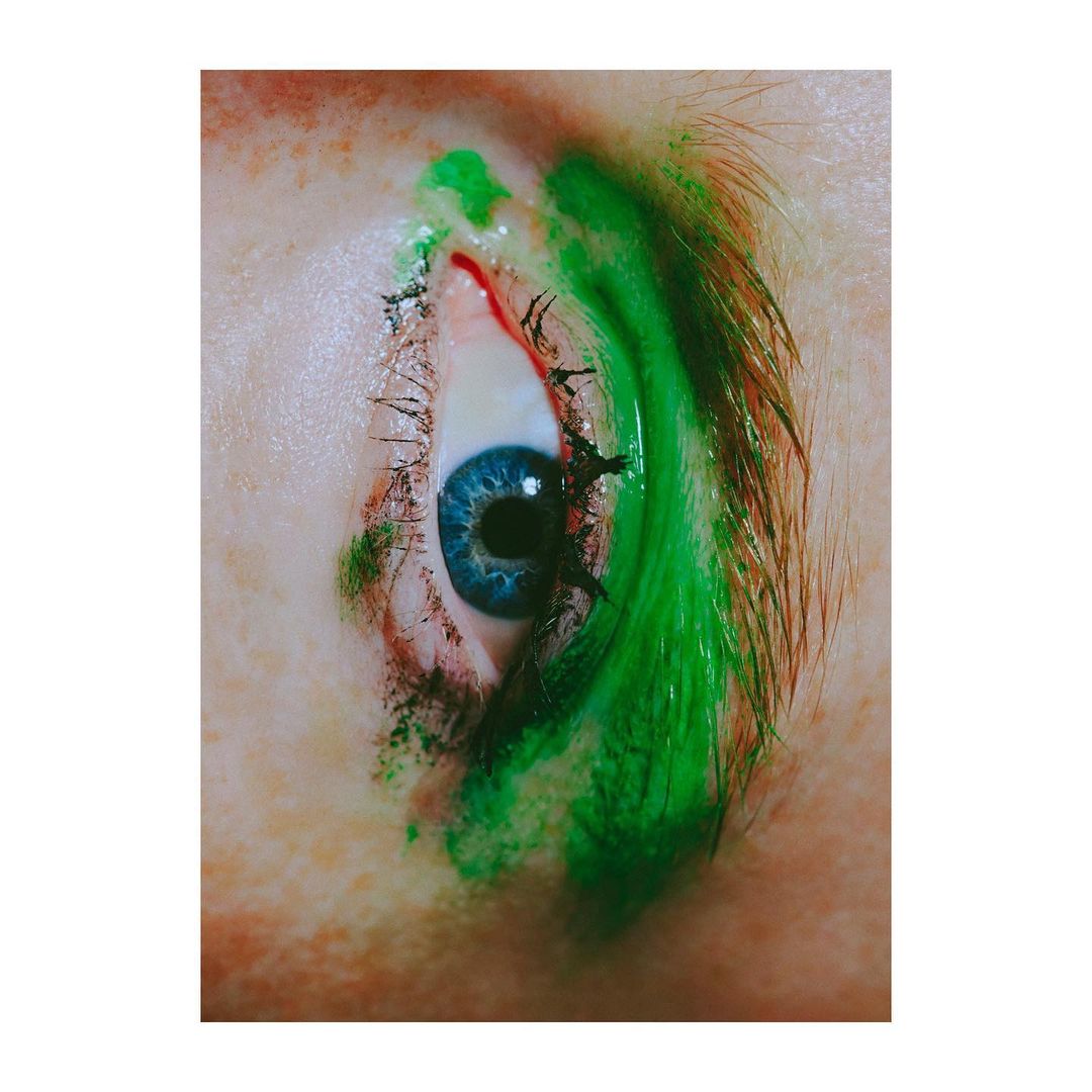 Close-up studio shot of a blue eye with green makeup around the eyelashes and eyebrow of an adult female with fair skin.
