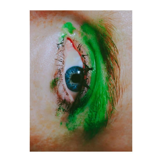 Close-up studio shot of a blue eye with green makeup around the eyelashes and eyebrow of an adult female with fair skin.