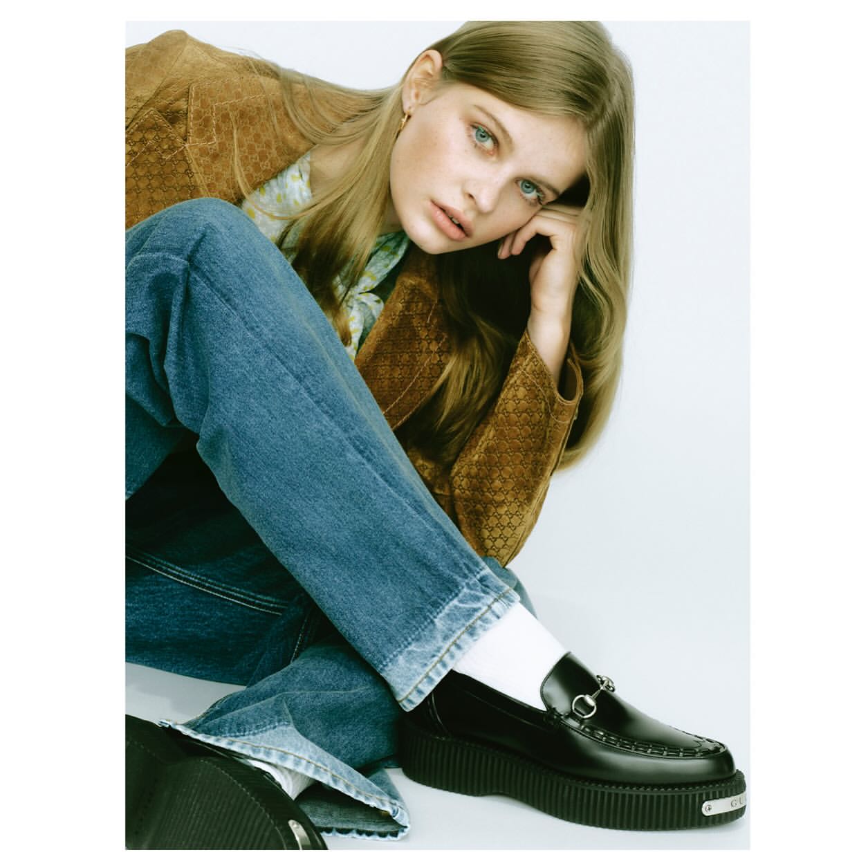 Young woman in a brown jacket and blue jeans seated against a plain white background