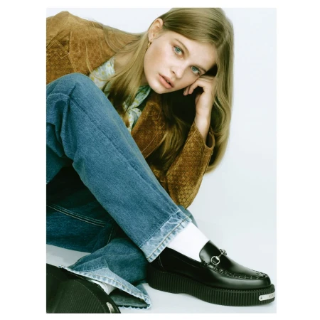 Young woman in a brown jacket and blue jeans seated against a plain white background