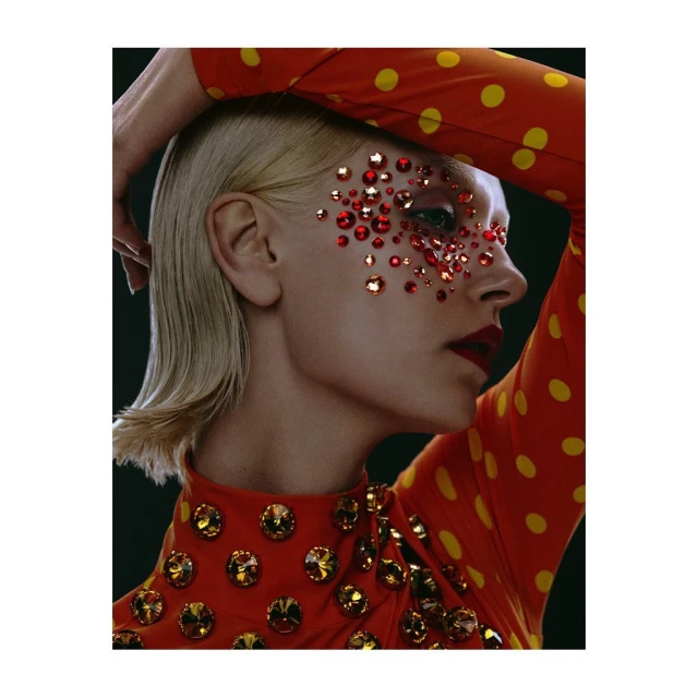 Close-up of a blond woman in an orange polka dot dress, adorned with red gemstones around her eyes, creating a striking beauty portrait.