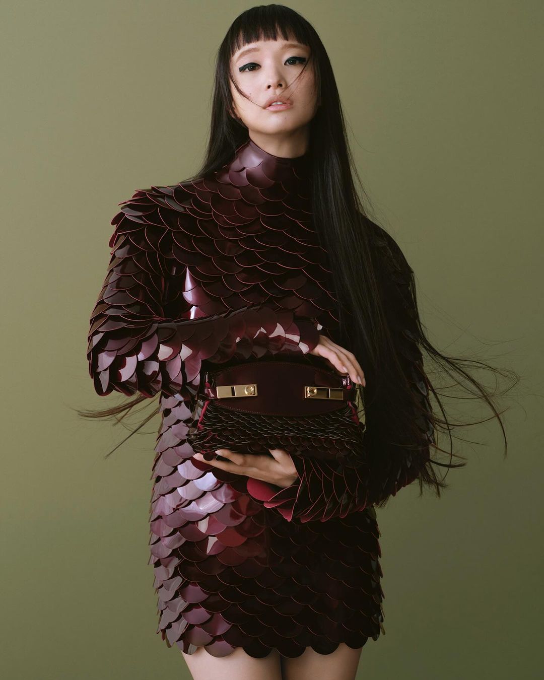 Portrait of a stylish Asian woman in a burgundy, scale-textured dress, holding a matching handbag against a muted green background.