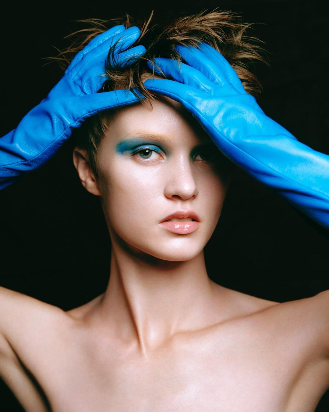 Close-up portrait of a woman with bare shoulders wearing blue eye makeup and blue gloves holding her spiky brown hair against a black background.