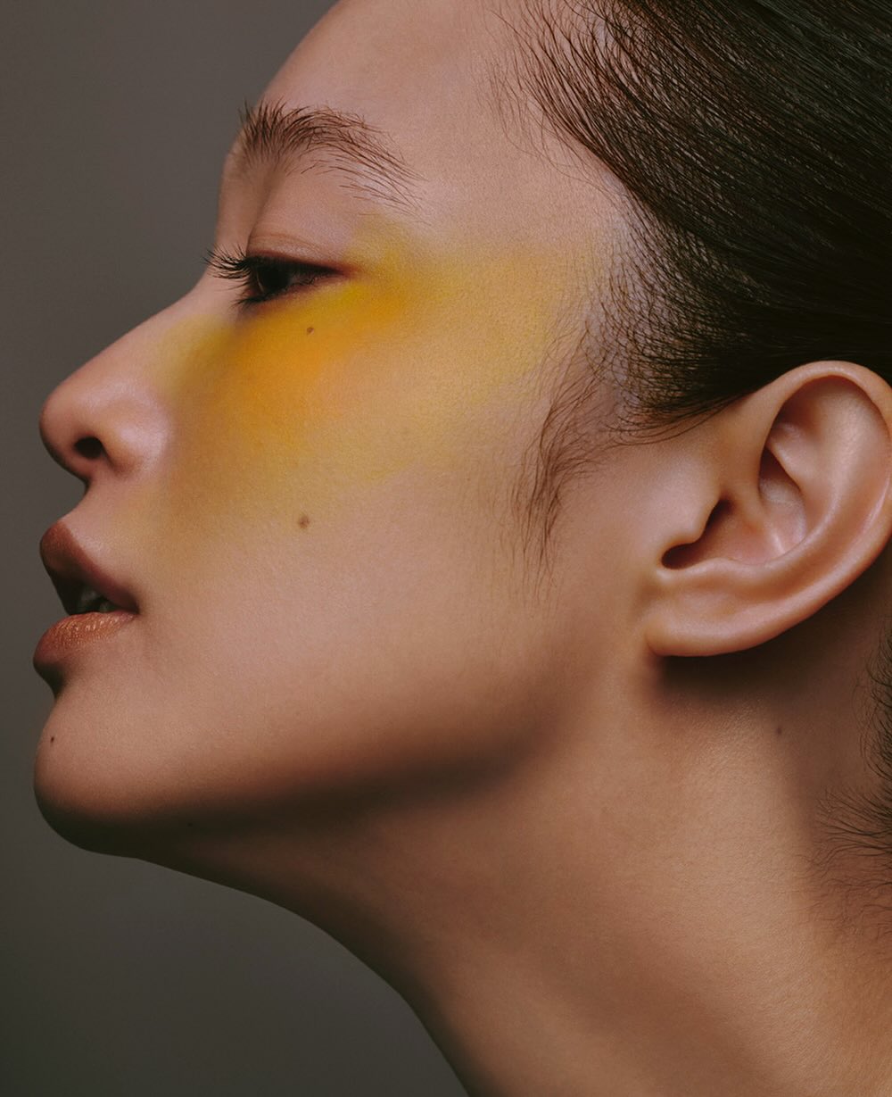 Asian adult female portrait with yellow makeup on face in profile, medium shot in photo studio against neutral gray background.