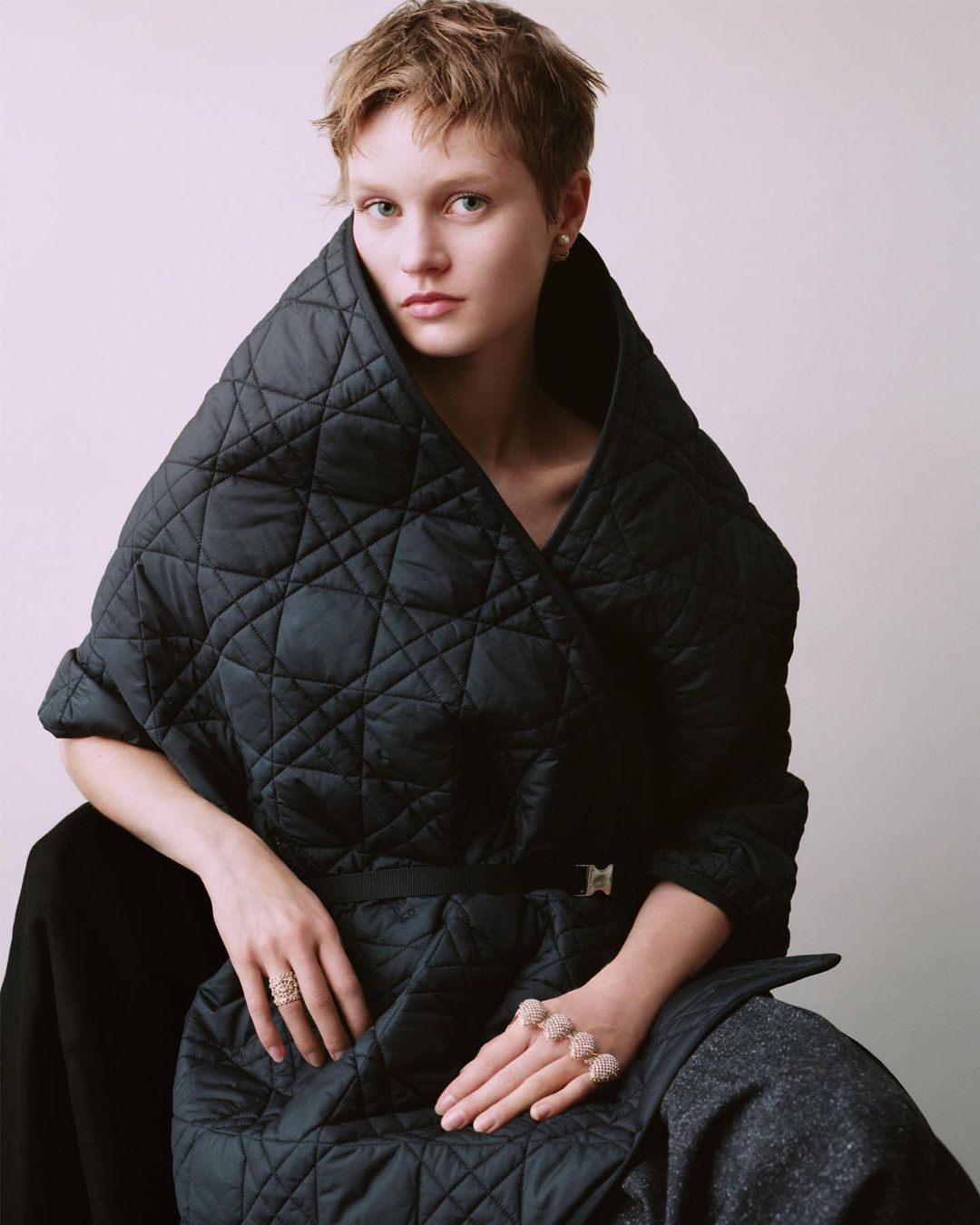 Elegant woman with short hair wearing a black quilted coat with a belt, seated indoors. The picture has soft, natural lighting.