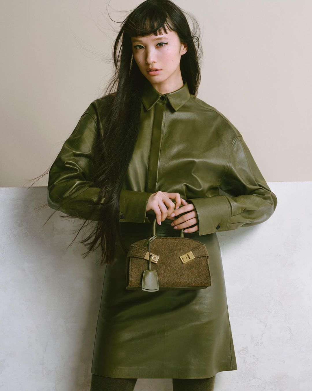 Asian woman in green leather dress and shirt holding brown tweed handbag in studio with soft lighting. Fashion and beauty photography.