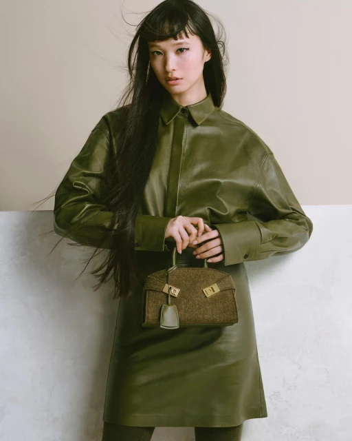 Asian woman in green leather dress and shirt holding brown tweed handbag in studio with soft lighting. Fashion and beauty photography.