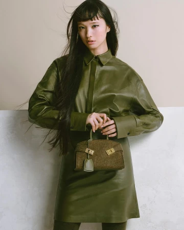 Asian woman in green leather dress and shirt holding brown tweed handbag in studio with soft lighting. Fashion and beauty photography.