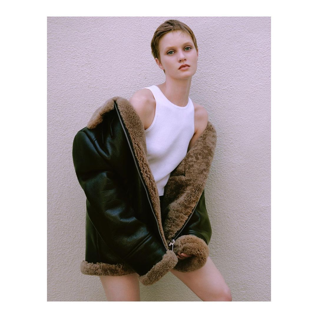 Woman poses in front of a neutral wall wearing a white shirt and oversized brown leather shearling coat in a fashion-style portrait.