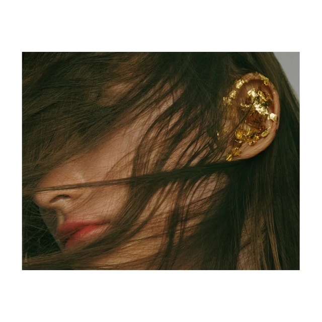 Close-up portrait of an adult woman with brown hair partially covering her face, adorned with gold leaf on her ear, set against a grey background.