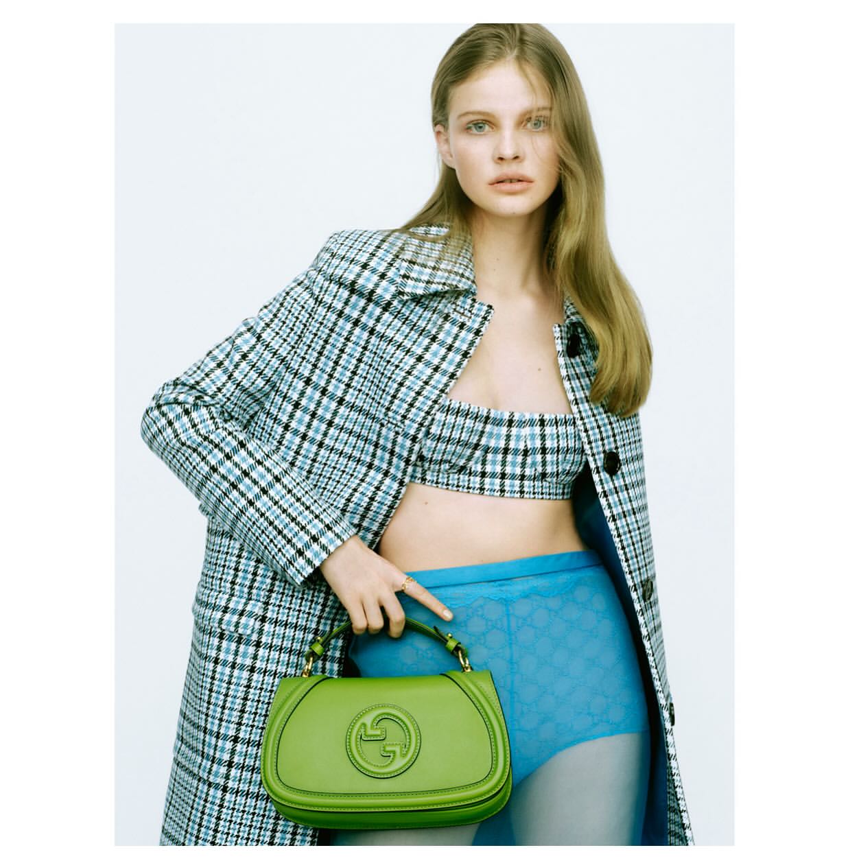 A fair-skinned woman poses in a blue plaid coat and bra top, holding a bright green Gucci purse, against a white background.