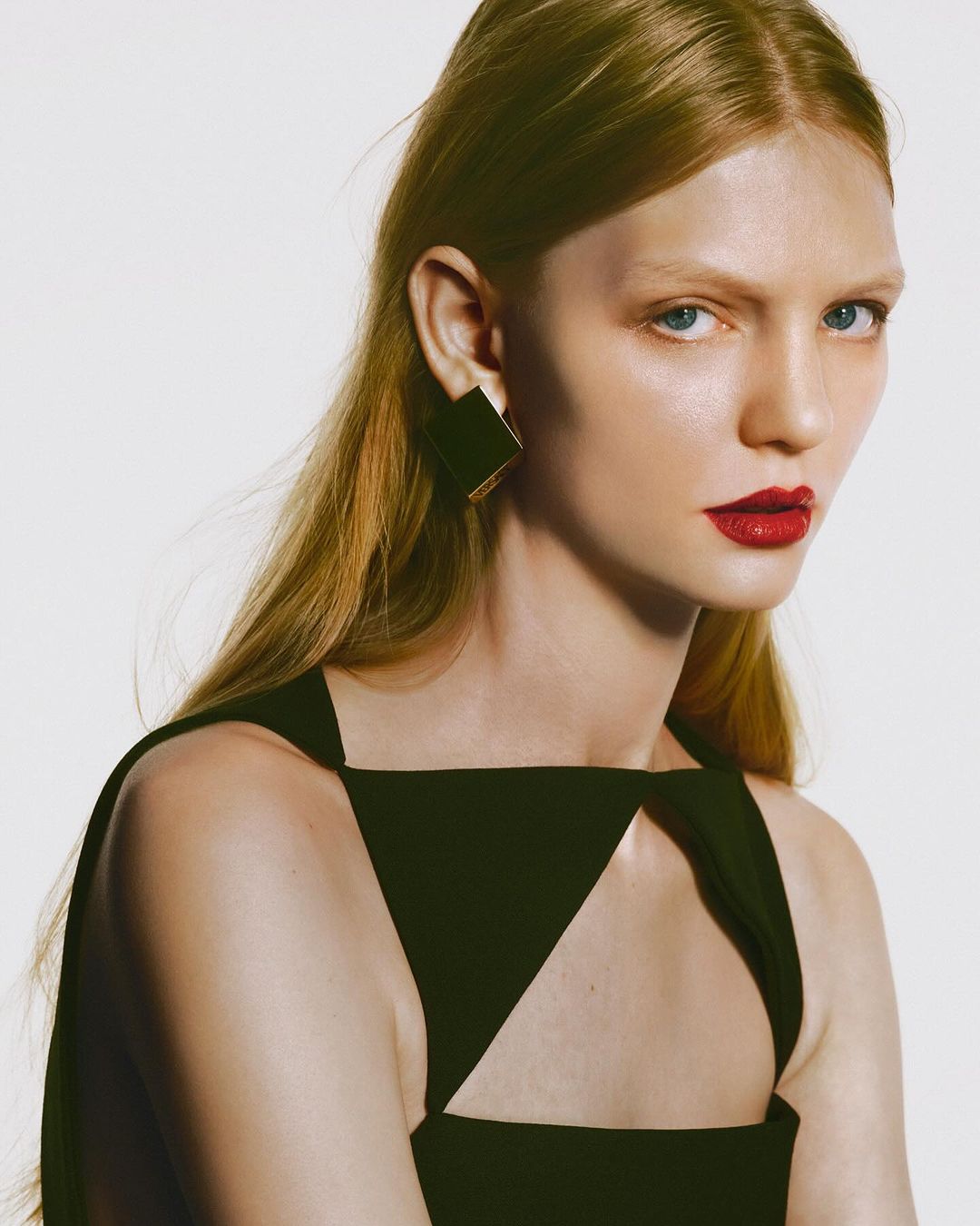 Close-up of an adult female fashion model with light hair, red lipstick, wearing a black top and square earrings in a photo studio.