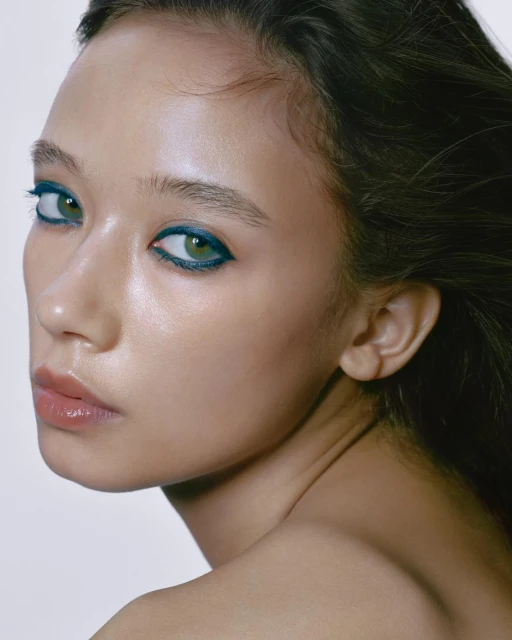 Close-up portrait of an adult female with green eyes and blue eyeliner, soft lighting, and a smooth skin tone against a white background.