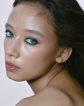Close-up portrait of an adult female with green eyes and blue eyeliner, soft lighting, and a smooth skin tone against a white background.