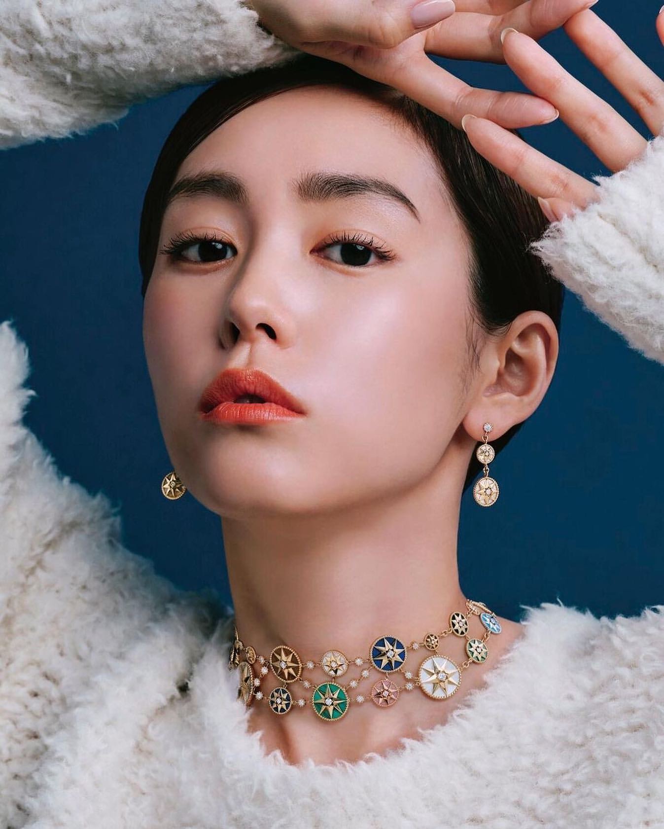 Close-up of a young Asian woman wearing an elaborate necklace and matching earrings against a dark blue background in a studio.