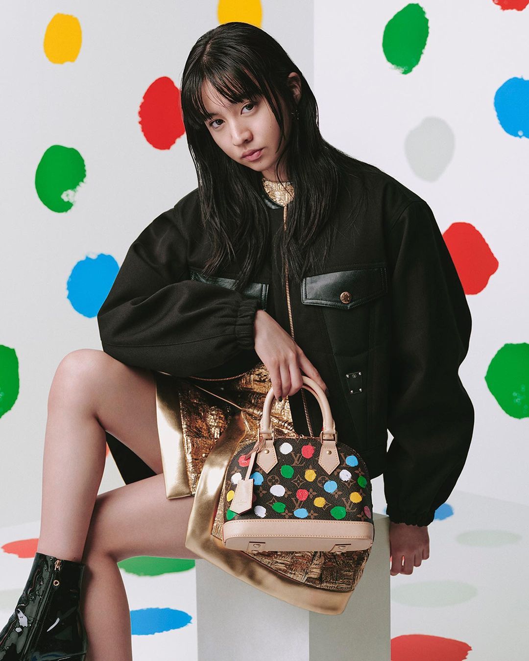 A woman holding a multicoloured polka dot handbag while wearing a black jacket and metallic gold skirt against a dot backdrop.