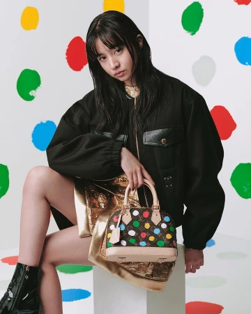 A woman holding a multicoloured polka dot handbag while wearing a black jacket and metallic gold skirt against a dot backdrop.