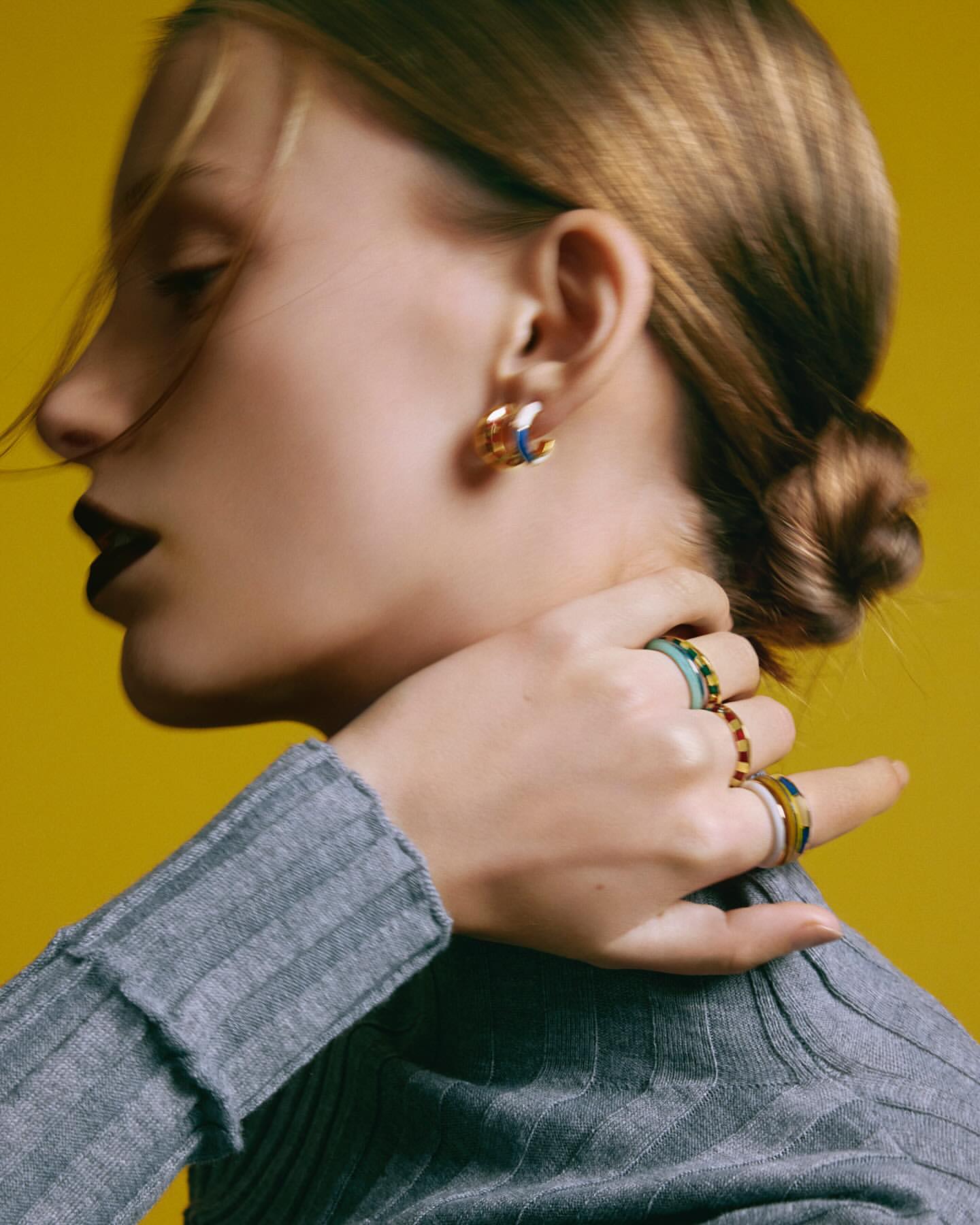 Profile portrait of a woman wearing jewellery and a grey ribbed turtleneck against a solid mustard yellow background.