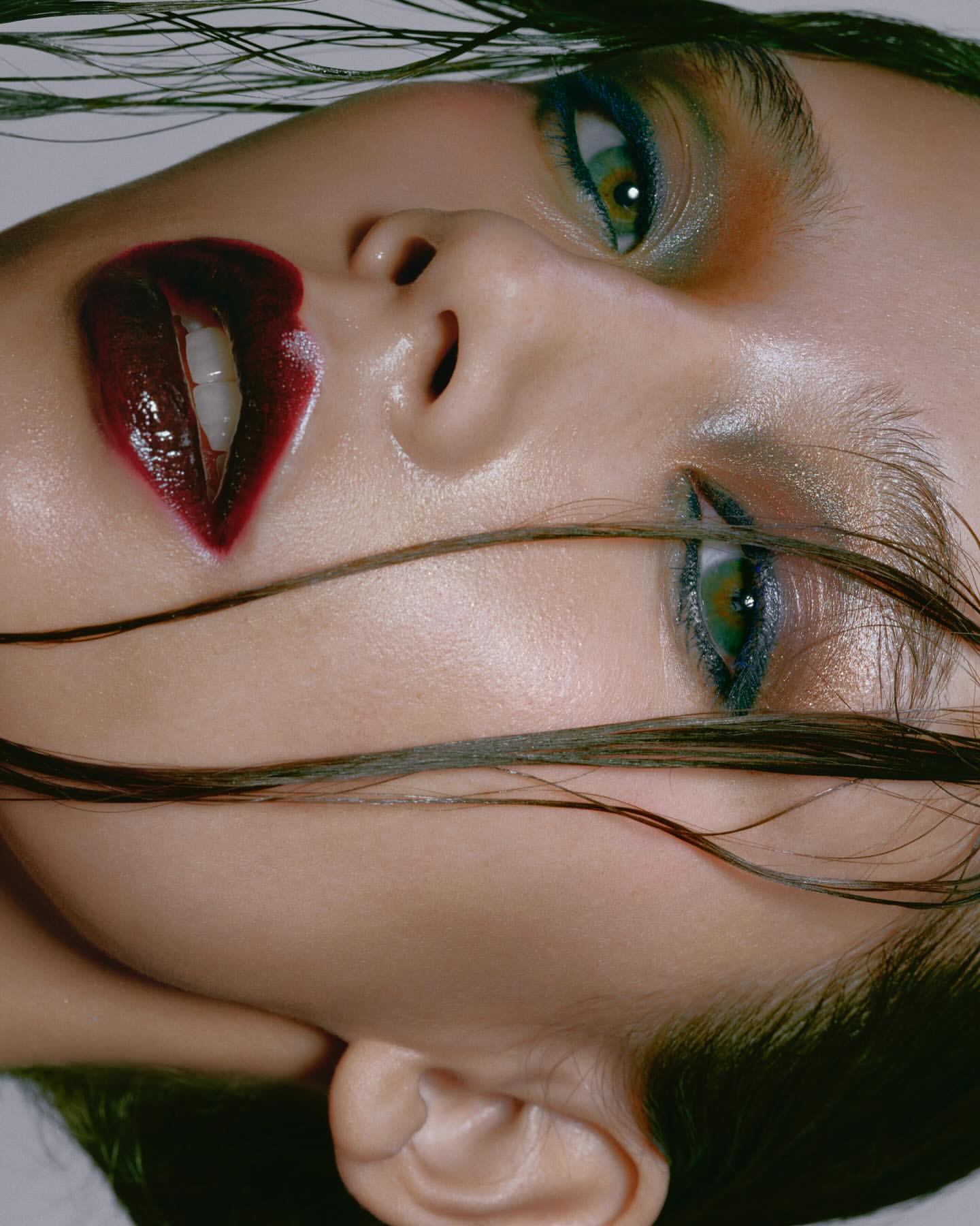 Close-up beauty portrait of a woman with wet hair, dark red lipstick, and green eyeshadow against a light background.