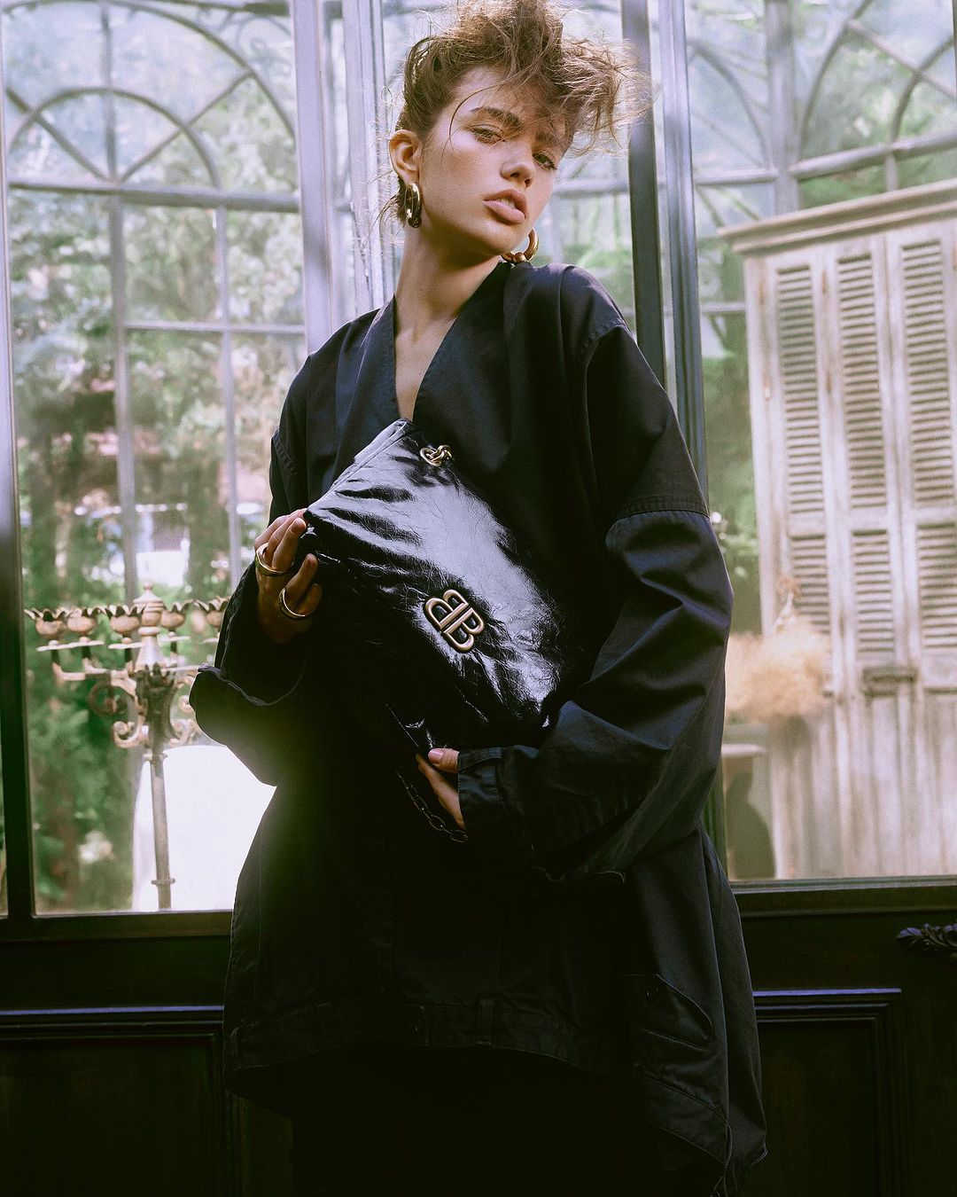 Adult woman poses with a Balenciaga handbag in an indoor setting with a greenhouse window backdrop, showcasing fashion and style.