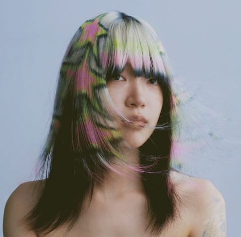 Close-up of an Asian woman with shoulder-length black hair that is blonde with colorful star patterns at the top, against a light blue backdrop.