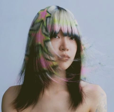 Close-up of an Asian woman with shoulder-length black hair that is blonde with colorful star patterns at the top, against a light blue backdrop.