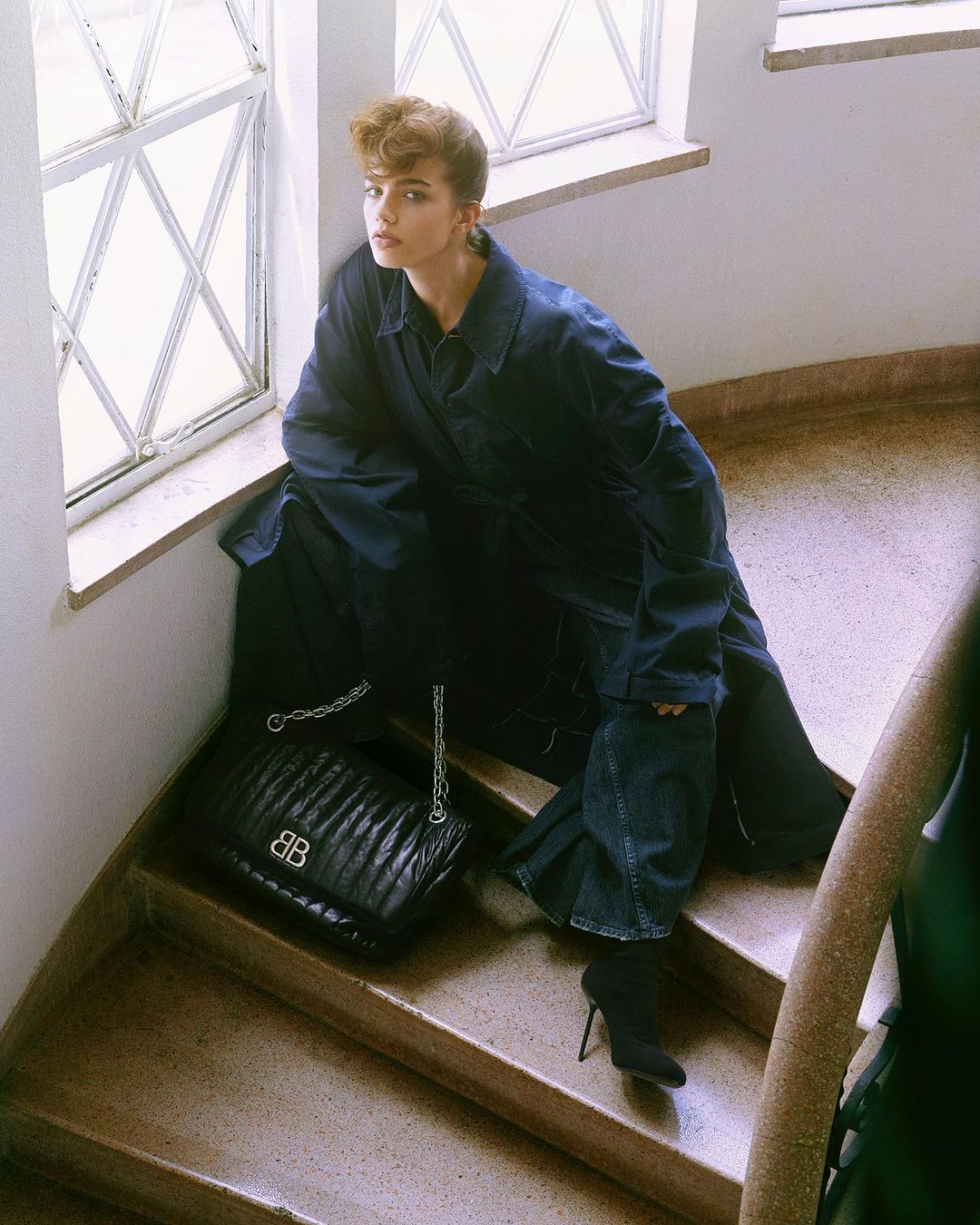 Woman in oversized denim jacket and stiletto boots poses on staircase next to black Balenciaga handbag and window with diamond panes.