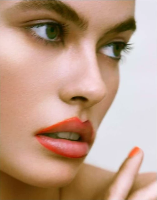 Close-up beauty shot of a woman with red lipstick and green eyes, face angled toward the upper right, and a clean, bright background.