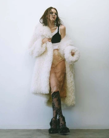 Model wearing a white furry coat over a black and white top against a plain wall.