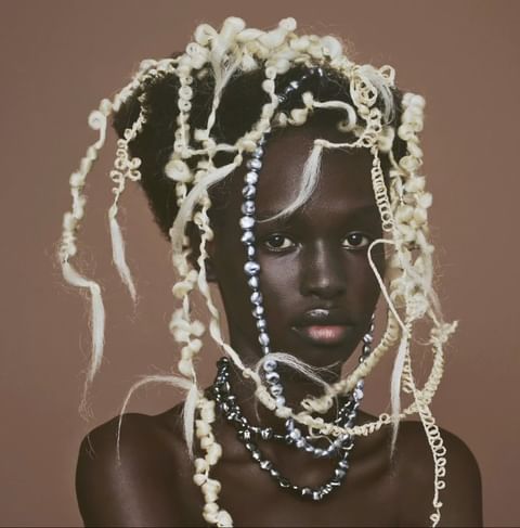 Portrait of a Black woman with sculptural white curled hair and layered pearl necklaces against a brown background.