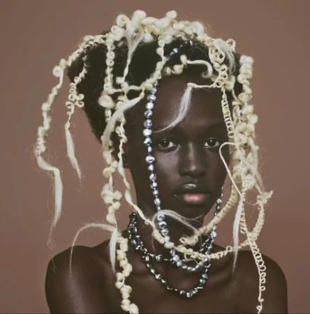 Portrait of a Black woman with sculptural white curled hair and layered pearl necklaces against a brown background.