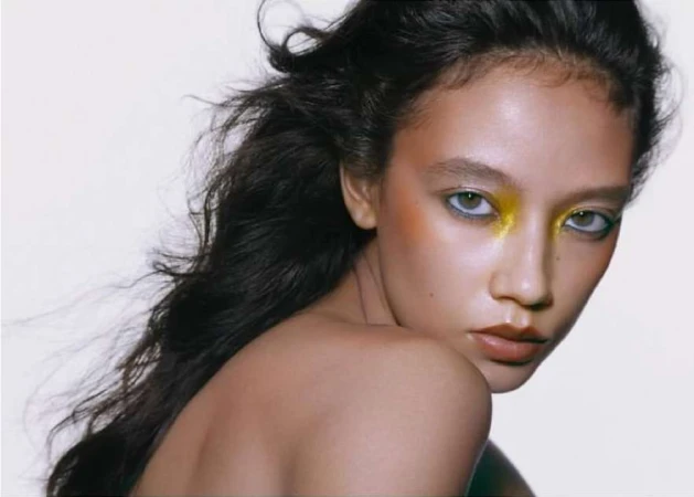 Close-up beauty portrait of a woman with vibrant yellow eyeshadow and dark tousled hair against a plain white background.