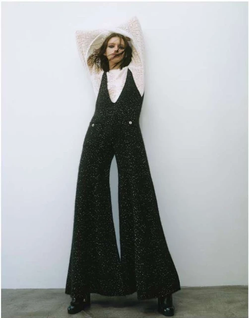 A woman poses in a studio, wearing a white sweater and black, glittery overalls with wide-legged pants and black shoes, against a white wall.
