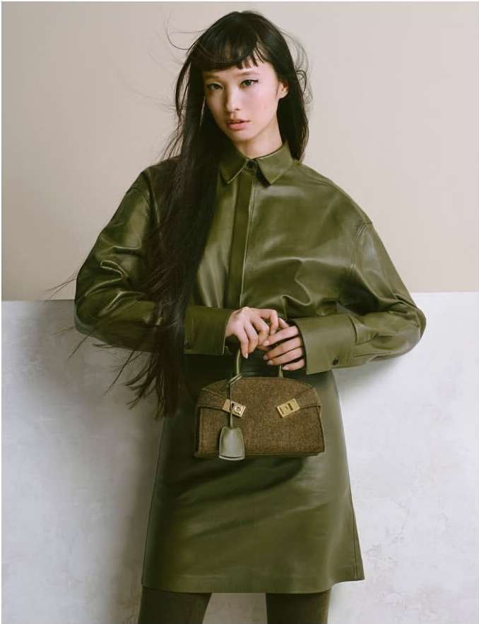 Asian model poses in an olive green, long-sleeved leather dress, holding a matching bag against a neutral two-tone background.